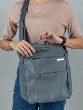 Kahverengi uzun kollu gömlekli ve kot pantolonlu kadın modelin yakın çekim stüdyo resmi gri arka planda uzun omuz kayışı olan modaya uygun gri renkli, çok amaçlı uniseks çantası asılı duruyor..