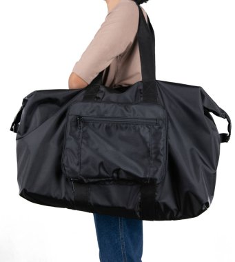 Uzun kollu kahverengi gömlekli ve kotlu kadın modelin izole edilmiş yakın çekim stüdyo fotoğrafı beyaz arka planda büyük siyah, çok amaçlı uniseks seyahat eden spor çantasının asılı duruşu..