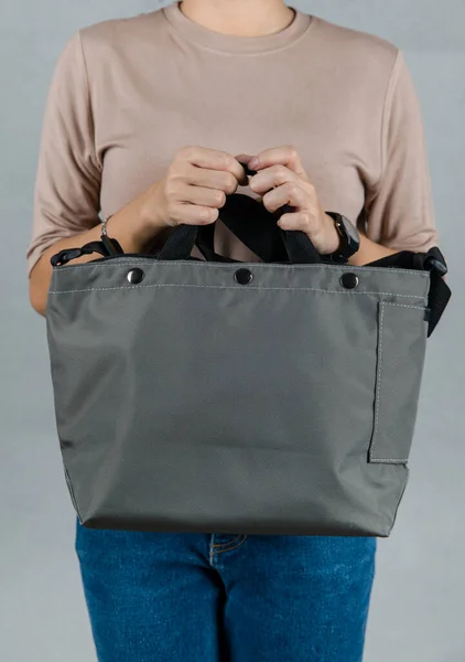 Closeup studio shot of female model in brown long sleeve shirt and jeans stand holding handle of fashionable trendy urban big blue unisex handbag with long shoulder strap in front of grey background.