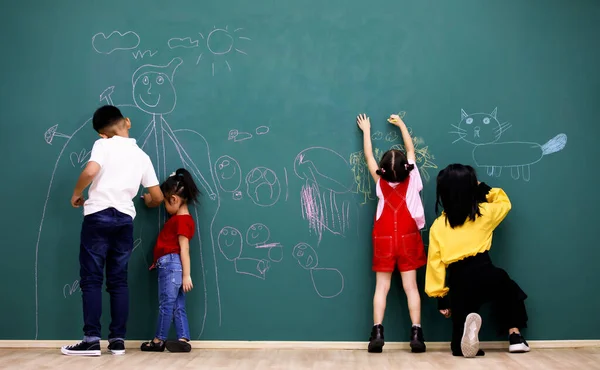 Asian little happy smart elementary primary schoolboy and schoolgirls students having fun together with friends using chalk writing drawing cartoon characters on green chalkboard in front classroom.