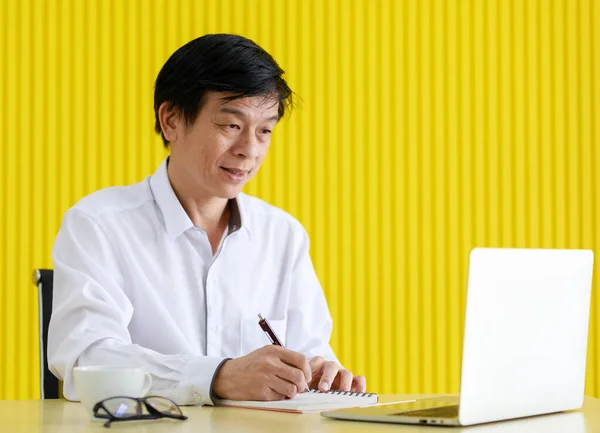 Asia old middle aged stress black hair male businessman wears white long sleeve shirt sit thinking looking at silver laptop on office working desk full of equipments in front yellow stripe background.