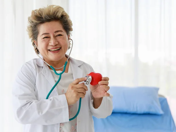 Portrait asian senior doctor elderly woman wearing uniform and holding stethoscope listening to internal sounds smiling look happy and comfortable standing near bed and look at camera in room.