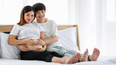 Yatakta birlikte oturan, beyaz gecelik giyen Asyalı genç bir adamın portresi. Mutlu bir şekilde gülümsüyorlar ve hamile bir annenin karnına dokunuyorlar..