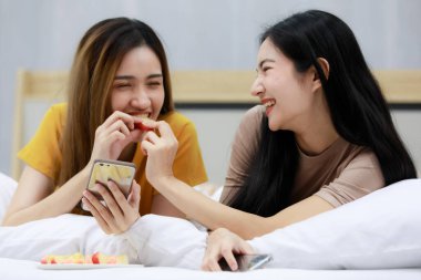 Gülümseyen iki Asyalı genç kadın beyaz yatakta dinleniyor ve mutlu elmalar besliyor. Sıcak bir yerde birlikte komik kadınlar. Aşık homoseksüel lezbiyen lgbt, lgbtq, lgbtq+