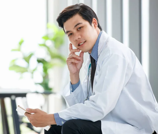 病院の医療室に座って携帯電話を閲覧する忙しいアジアの男性開業医の側面図 — ストック写真