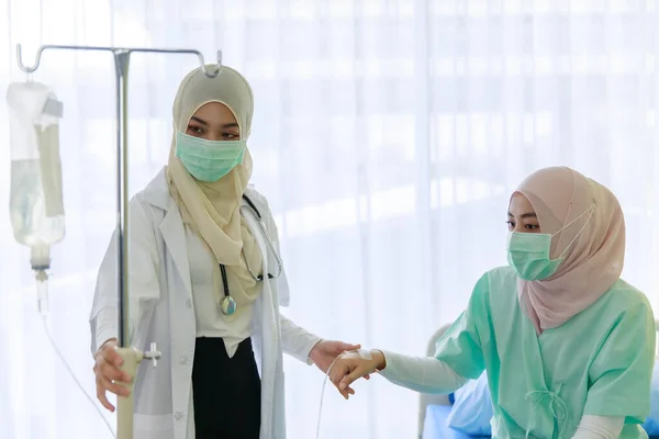 若い女性のイスラム教徒の医師は 病院の部屋で患者を回復するために使用される生理食塩水の袋のチューブを保持しながら イスラム教徒の患者の健康をチェックフェイスマスクを着用 Covid 19パンデミックの概念 — ストック写真