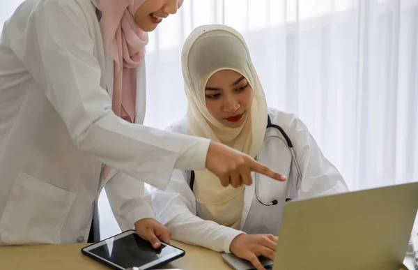 Two young female Muslim doctors are working and discussing using laptop and tablet at the hospital office room.