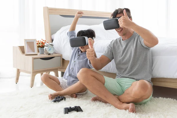 Father and his mixed-race son wearing vr virtual reality device to play a game together with fun and happy in bedroom