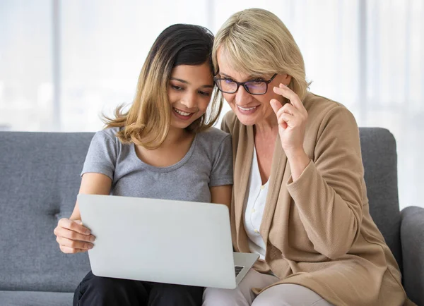 Positive young Asian female explaining how to find information on internet to middle aged mother in law while sitting together on sofa at home