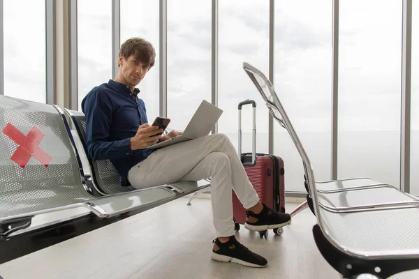 Male tourist sitting on row seats in airport lobby chair with social distancing sign and using laptop notebook computer and smartphone connect to internet for work . New normal journey concept.