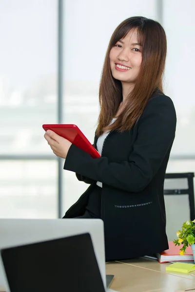 身穿黑色西服的年轻迷人的亚洲女人坐在桌子上 手持红色的平板电脑 自信地看着办公室 窗户背景模糊不清 现代办公室生活方式的概念 — 图库照片