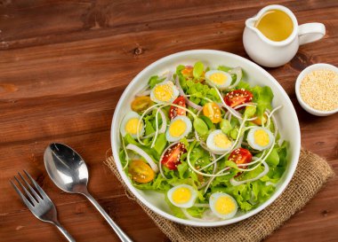 Büyük beyaz bir kase marul, haşlanmış yumurta, soğan ve domates salatası ile Japon susam yağı sosunu ahşap masa üzerinde küçük zeytinyağı kavanozu ve susam kabı ile karıştırın..