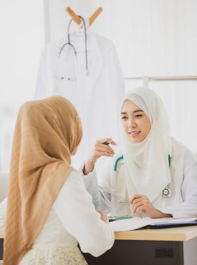 Çekici, gülümseyen, kahverengi tesettürlü Müslüman kadın masada oturuyor, bakıyor ve doktorla konuşuyor. Bulanık doktorlu bir hastaya seçici odaklanma.