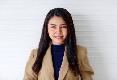 Çekici gülümseyen, kahverengi takım elbiseli ve mavi elbiseli genç bir Asyalı kadının, stüdyoda beyaz arka planda izole edilmiş kameraya bakarken, yatay yakın plan portresi.