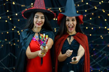 Şık cadı kıyafetleri ve şapkaları içinde neşeli ve yılanlı krakerleri Cadılar Bayramı partisinde kameraya çeken neşeli genç bayan arkadaşlar.