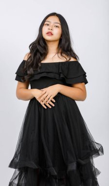 Siyah elbiseli sevimli, gülümseyen bir genç kızın dikey portresi. Stüdyoda beyaz arka planda kameraya bakıyor. Yeni kız şık kıyafetler içinde utangaç duygularla saçlara dokunuyor.