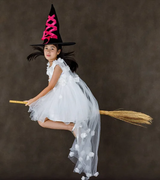 ポートレートスタジオショットの小さなかわいいアジアの子供でピンクの魔女の衣装で黒の高い帽子の表情でカメラポーズ飛行ジェスチャー乗って魔法の魔女ブロムスティック上のハロウィーンの伝統的な祭り — ストック写真