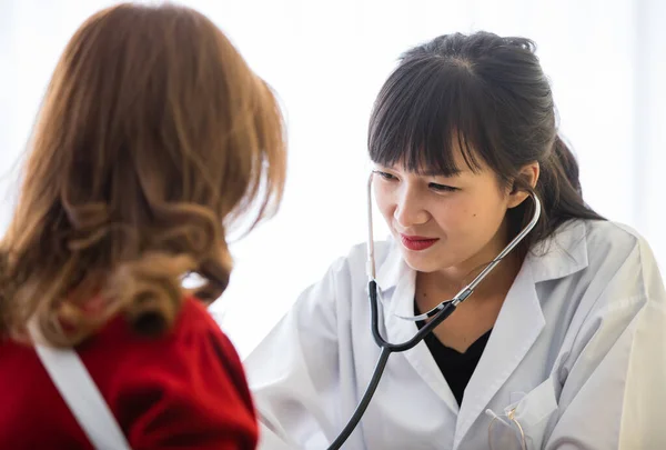 Doctor with long black hair white lab coat sitting checking heartbeat of lady who has blond hair wear red shirt in hospital healthcare center before discussing for recovering process