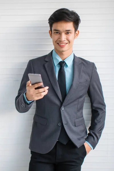 灰色の壁に立っている間スーツの笑顔とブラウジング携帯電話で幸せな若いアジアのマネージャー — ストック写真