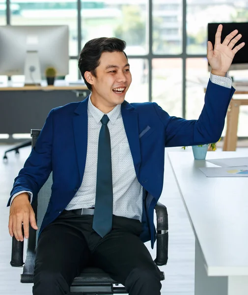 Young and elegance Asian businessman in blue suit sitting in modern office and rising hand greeting or say hello to someone.