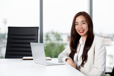 Başarılı, Asyalı, zarif takım elbiseli, başarılı bir iş kadını. Bilgisayarlı masada oturmuş modern ofiste çalışırken kameraya bakıyor..