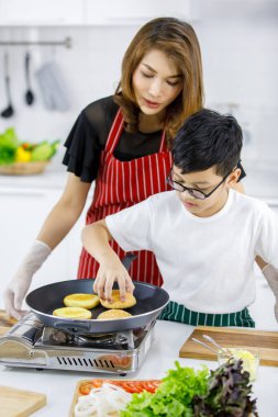 Asyalı oğul, mutfakta hamburgerleri pişirirken annesinin yanında sıcak çörekleri sıkıştırıyor.
