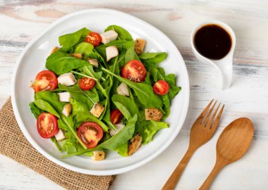 Domatesli taze ıspanak salatası, kahverengi çuval bezinde beyaz seramik tabakta kızartılmış küçük sebzeli tavuk parçaları, balzamik sirke ve beyaz ağaçlık masada çatal bıçak. Sağlıklı kavram.