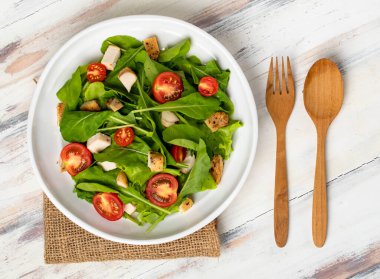 Fresh spinach salad with tomato, small pieces of herb roasted chicken in white ceramic dish on brown sack cloth on white wooded table. Concept for healthy. clipart