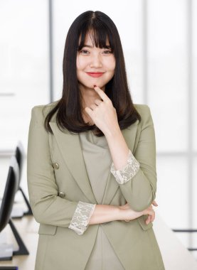 Çekici gülümseyen Asyalı bir kadının dikey portresi. Uzun kollu bir elbise içinde masada oturmuş kameraya bakarken. Ofiste poz veren genç profesyonel bir kadın.