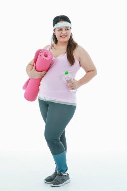 Asyalı mutlu, sağlıklı, spor giysili, kafa bandı takan, gülümseyen, beyaz arka planda elinde pembe yoga minderiyle su şişesi tutan şişman şişman bir kızın izole edilmiş bir fotoğrafı..