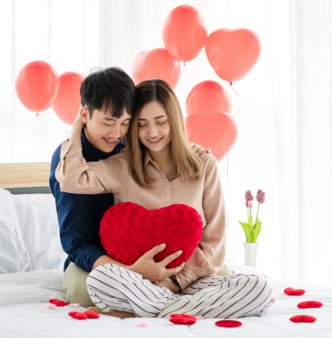 Yastık şeklinde kalp şeklinde gülümseyen ve yatakta birbirini kucaklayan iyimser genç Asyalı çift. Genç aşıkların aşk ve ilişkisi.