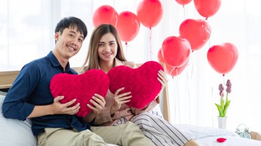 Kalp şeklinde yastıkları olan neşeli Asyalı erkek ve kadın aşıklar Aziz Sevgililer Günü 'nde yatakta dinlenirken gülümseyip kameraya bakıyorlar.