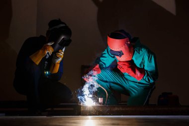 Gece vakti fabrika atölyesinde çalışan bir mühendis. Mekanik tulum ve eldiven giyen iki tamirci kaynaklama makinesiyle metal işçiliği yapan bir kaynak kaskı taktı..