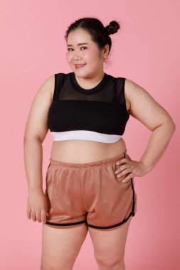 Şort ve spor elbiseli, çekici, iri vücutlu, sportif görünümlü bir kız ayakta duruyor ve kendine güvenerek gülümsüyor. Yüzü ve duyguları olumlu. Mutlu ve özgüven kavramı.