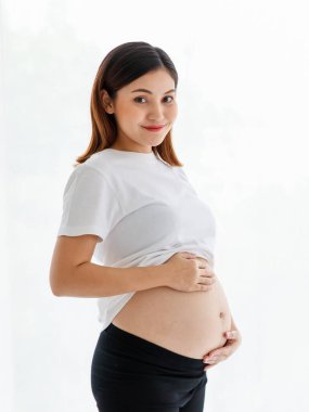 Gülümseyen genç, mutlu, hamile bir Asyalı kadın, gelişigüzel ayakta durup karnına sevgiyle dokunuyor ve beyaz arka planda çekim yapabilmek için kameraya bakıyor..