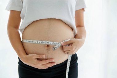 Güzel, mutlu, Asyalı hamile bir kadının portresi gülümseyerek ayakta dururken kameraya bakarken zili ölçer..