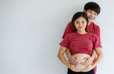 Gülen genç karı ve koca beyaz arka planda yan yana duruyorlar. Bir erkek hamile bir kadını kucaklar, elini karnına koyar. İnsanlar, aile ve yaşam tarzı konsepti. Boşluğu kopyala.