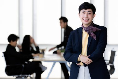 LGBTQ transseksüel ofis çalışanının portresi dikilirken ve hoş geldin jesti yaparken arka planda iş arkadaşları bulanıklaşır..