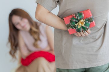 Asyalı, uzun kahverengi saçlı, kolsuz bir kadın turuncu koltukta oturuyor, yüzünü kırmızı yastığa koyuyor ve kameraya bakıyor. Erkek arkadaşı arkasında yeşil kurdele olan kırmızı hediye kutusunu saklıyor..