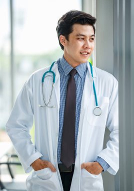 Üniformalı, neşeli, genç ve yakışıklı Asyalı erkek doktor. Steteskopu var, elleri klinikte duruyor ve dışarıya bakıyor.