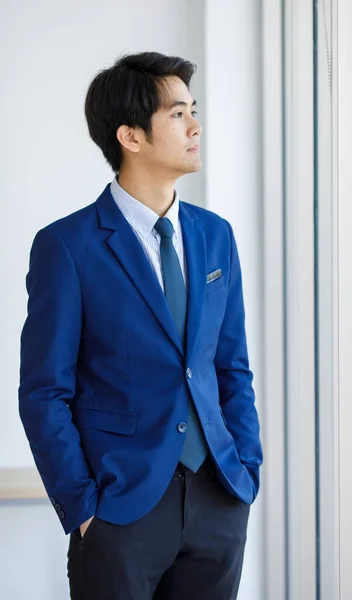 Portrait closeup shot of Asian young handsome happy intelligence college teen student wears blue formal suit and necktie standing hold hands in pants pockets look outside in front of white wall.