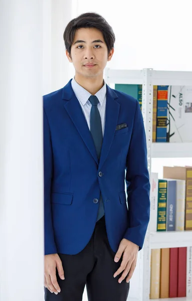 アジアの若いフレンドリーなスマート法学生のポートレートショットは 大学図書館で本でいっぱいのフロント本棚のカメラで青い正式なスーツとネクタイスタンド傾いた壁の表情を身に着けています — ストック写真