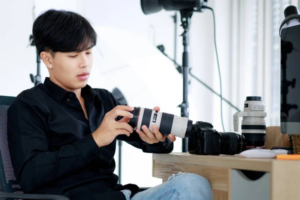 アジアのハンサムな男性写真家は カジュアルな黒のシャツを着て カメラを持ち ポーズをとって 軽い装備でスタジオに座っています — ストック写真