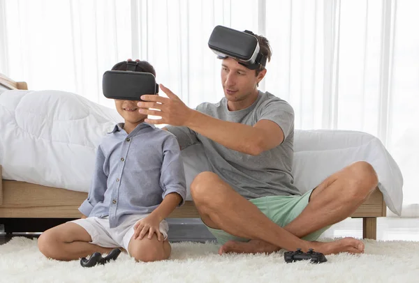Father and his mixed-race son wearing vr virtual reality device to play a game together with fun and happy in bedroom