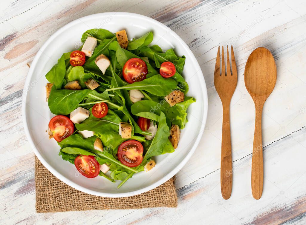 Fresh spinach salad with tomato, small pieces of herb roasted chicken in white ceramic dish on brown sack cloth on white wooded table. Concept for healthy.