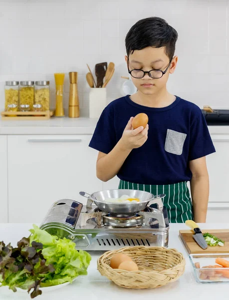 Smart kid standing in front of flying pan on gas stove in modern home kitchen looking at egg in hand as taking family cooking education from mom by learning to do healthy food of flied egg