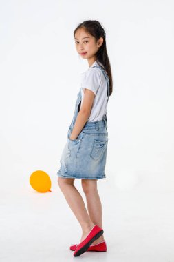 Portre, izole edilmiş, uzun siyah saçlı, kot etekli ilkokul kızının beyaz arka plan önünde kameraya gülümseyen fotoğrafına yakın..