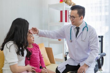 Gözlüklü genç Asyalı adam steteskopla gıdaklıyor ve annesinin yanında oturan küçük kızın alnına dokunarak muayene ediyor. Çocuk ve aile sağlık hizmeti kavramı.