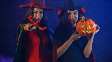 Cadı kostümlü, şapkalı şeytan mızrağı ve balkabağı kafası tutan ve Cadılar Bayramı partisinde kameraya bakan genç kızlar.