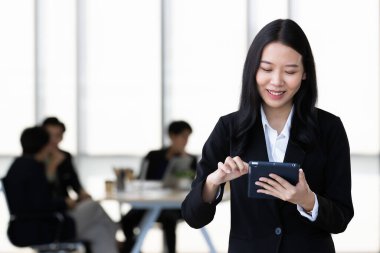 Siyah takım elbiseli genç Asyalı iş kadını elinde tablet bilgisayarla kameraya poz veriyor. Ekibi de arka planda bulanık konuşuyor..